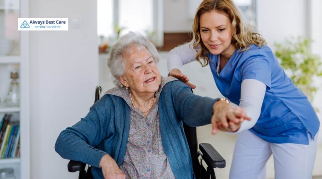 A caregiver gently helps an elderly woman stretch her arm in a living room setting. The woman is seated in a wheelchair, and the caregiver, in blue scrubs, is offering physical support with a caring smile.