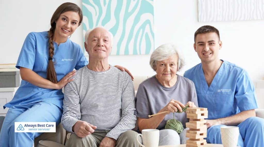 wo caregivers and two seniors engage in a fun and interactive activity at home. One caregiver stands beside a man, while the other sits beside a woman, both of whom are playing a game together. The atmosphere is relaxed and joyful.
