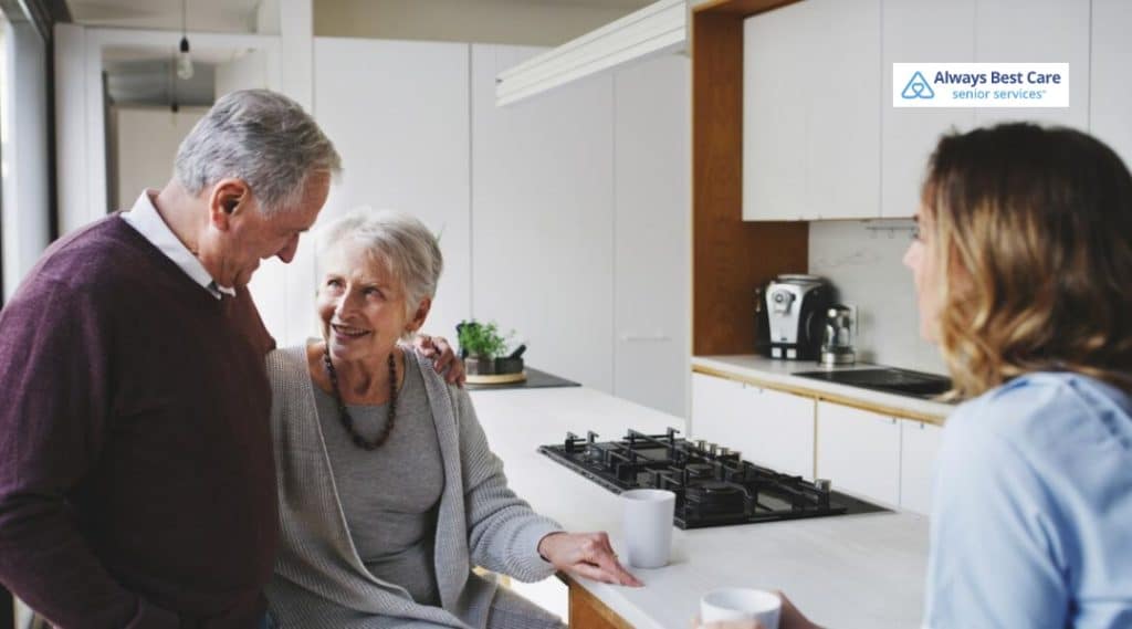miling older couple chats with a caregiver in a bright kitchen over coffee.
