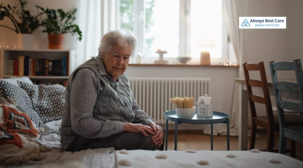 Older woman sitting on the edge of a bed in a bright bedroom with a small table nearby