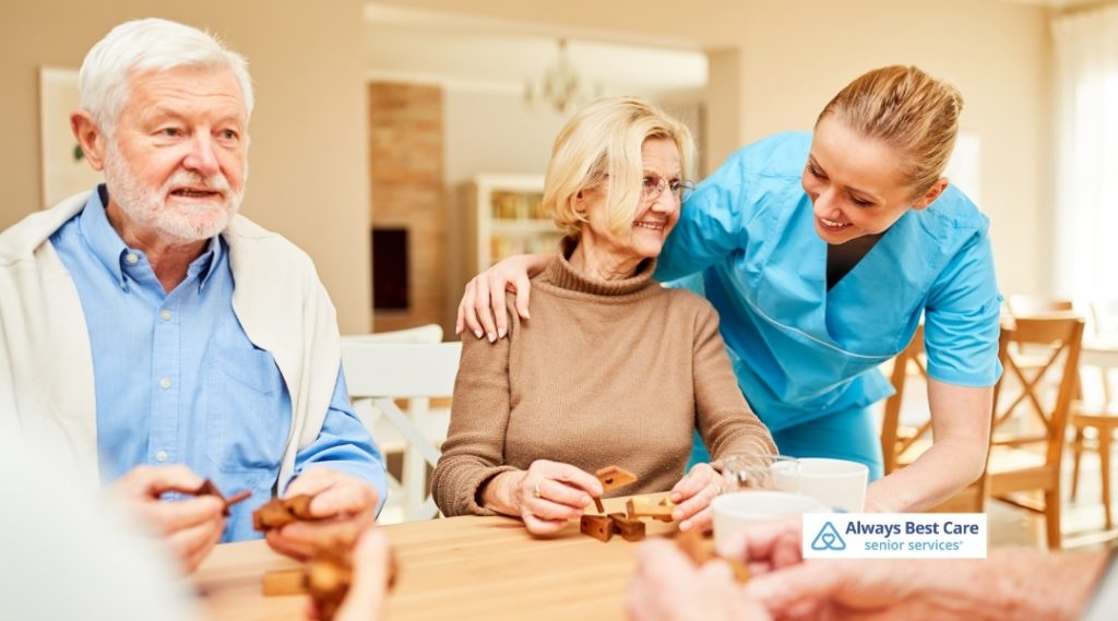 A cheerful caregiver in scrubs interacts with a smiling elderly woman while other seniors participate in a group activity. The Always Best Care logo is visible in the bottom right corner.