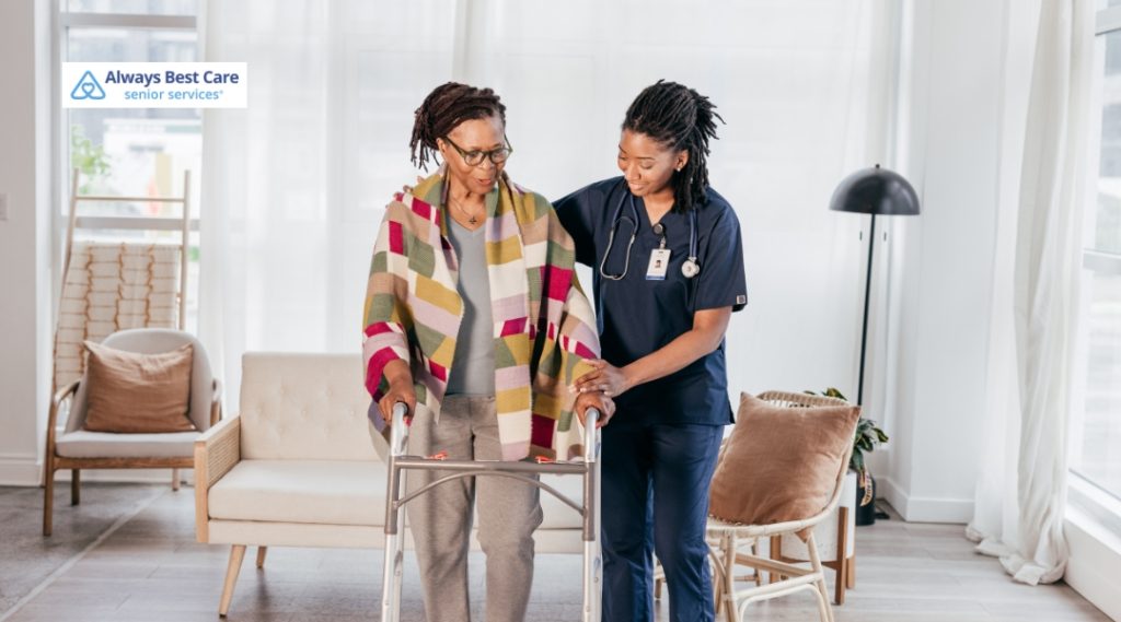A caregiver in navy scrubs is helping a senior woman with a walker in a bright, modern living room. The senior woman wears a colorful blanket and looks supported as she moves with the caregiver's help.