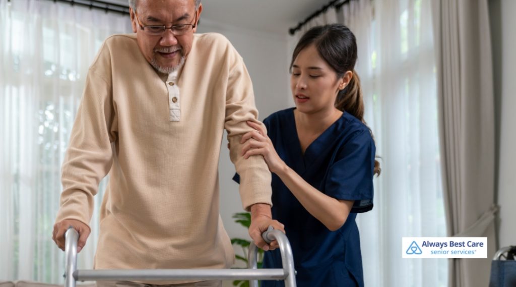 A caregiver in navy scrubs is assisting an elderly man with walking using a walker in a well-lit, comfortable home. The caregiver provides support by holding onto his arm as he moves forward.