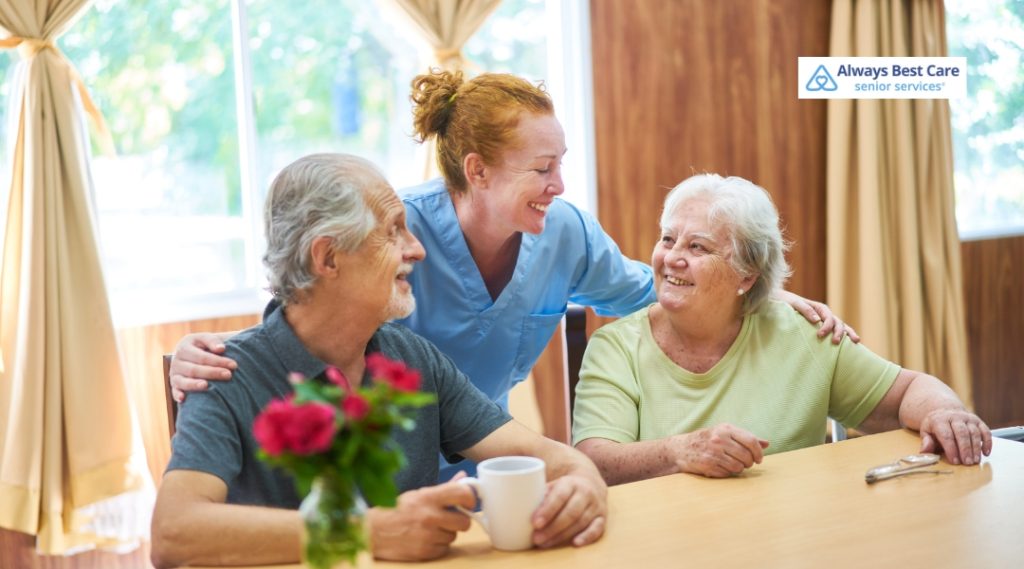 A caregiver in light blue scrubs is smiling and interacting with two senior clients sitting at a table. One elderly man and one elderly woman smile back, holding cups of tea or coffee, in a warm, inviting setting.