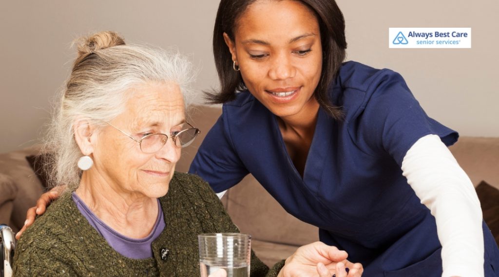 A senior woman with glasses looks intently at a glass of water as her caregiver, dressed in a blue uniform, assists her with a thoughtful, caring demeanor.