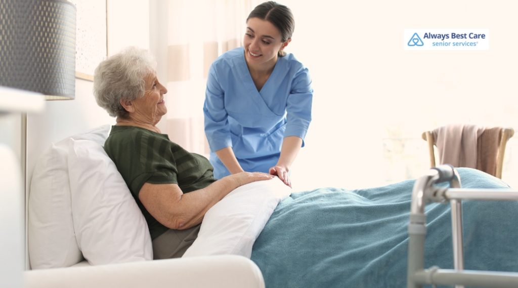 A smiling caregiver in blue scrubs assists an elderly woman resting in bed, offering comfort and support at home.