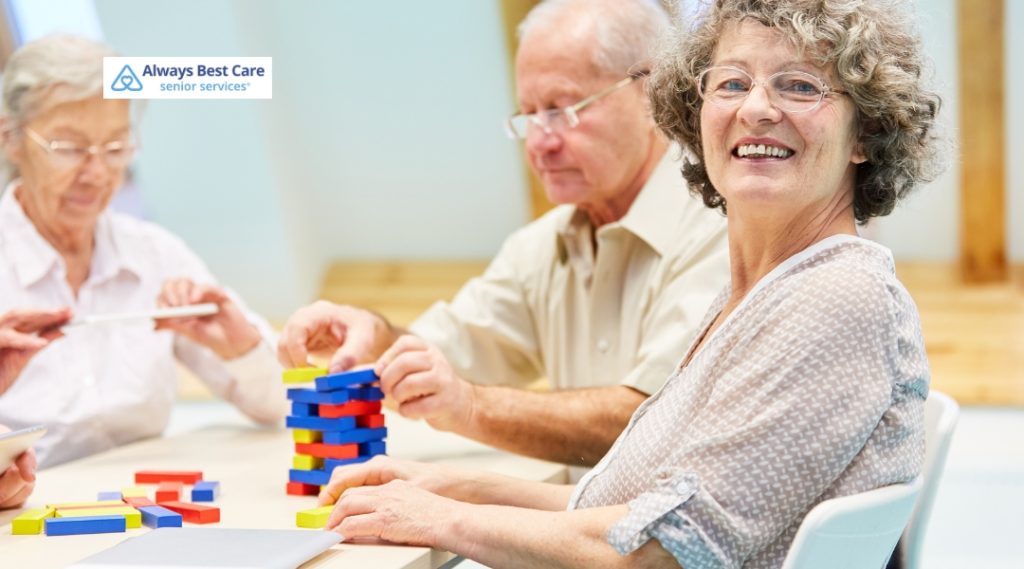 Three elderly individuals engage in a colorful block-stacking game at a table. One woman smiles at the camera, exuding joy and social interaction. The Always Best Care logo appears in the top left corner.