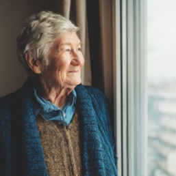 SENIOR WOMAN LOOKING OUT A WINDOW WITH SOFT NATURAL LIGHT, REFLECTING ON CONNECTION AND SUPPORT IN FITCHBURG, WI.
