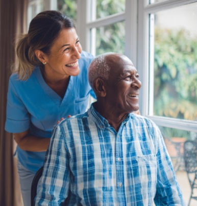 CAREGIVER SMILING WITH A SENIOR MAN BY A WINDOW, SHOWING COMPANIONSHIP AND SUPPORTIVE HOME CARE IN MADISON, WI. 