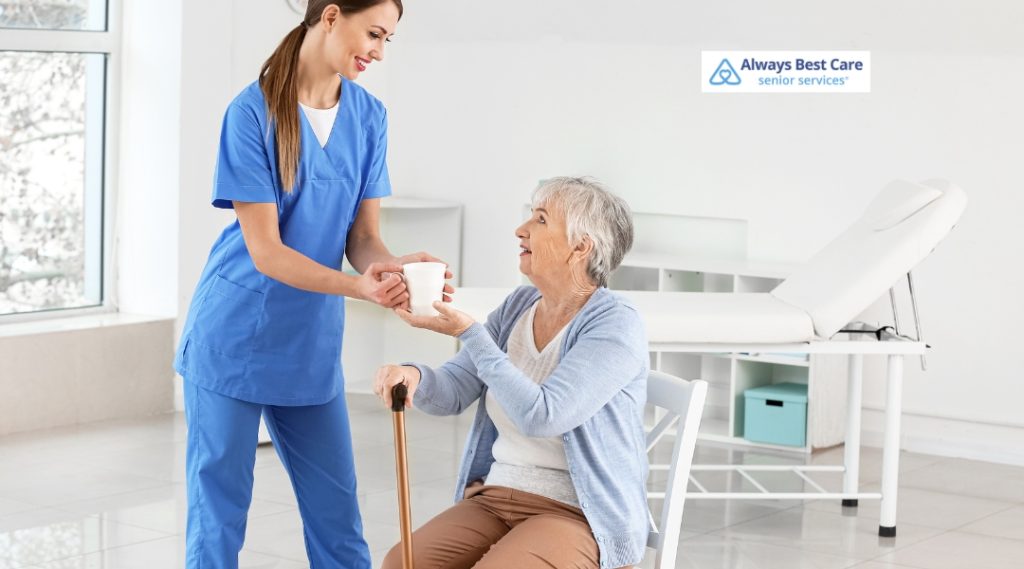 A caregiver hands a warm cup to an elderly woman sitting with a cane, as they smile at each other in a bright, minimalistic indoor space, emphasizing compassion and care.