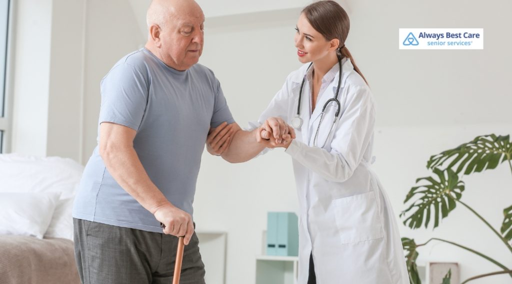 A senior man, using a cane, is assisted by a female healthcare professional in a white coat as he stands. The scene is set in a modern, well-lit home with a comforting ambiance.