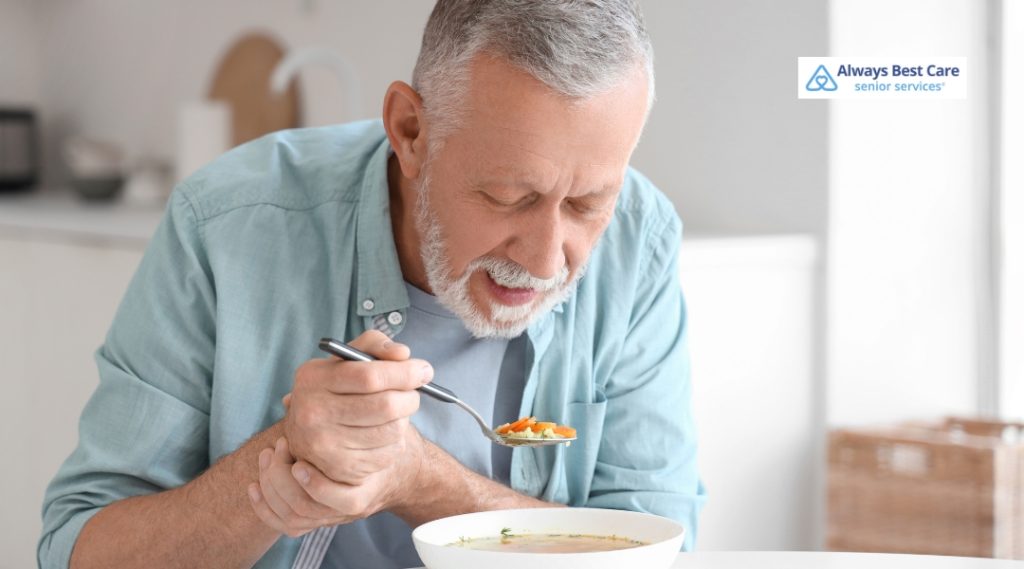 A senior man with gray hair is smiling while holding a spoon with soup, seated at a table in a cozy, modern kitchen. He appears to be enjoying his meal in a calm and comforting environment.
