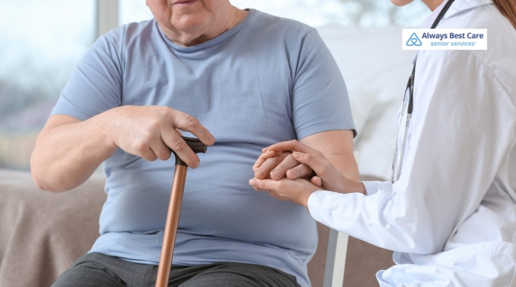 A senior man, dressed in a blue t-shirt, is holding a walking cane with one hand while a healthcare worker, wearing a white coat, gently holds his hand to assist him. The setting is a bright and peaceful home.