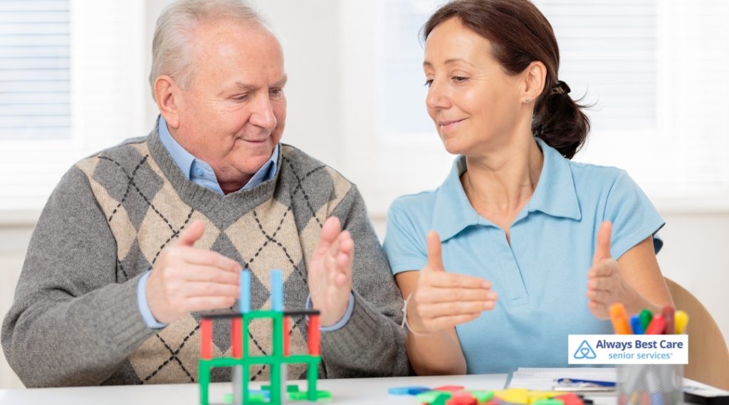 A senior man working with a caregiver, stacking colorful building blocks as part of a cognitive activity in a bright, cheerful indoor space.