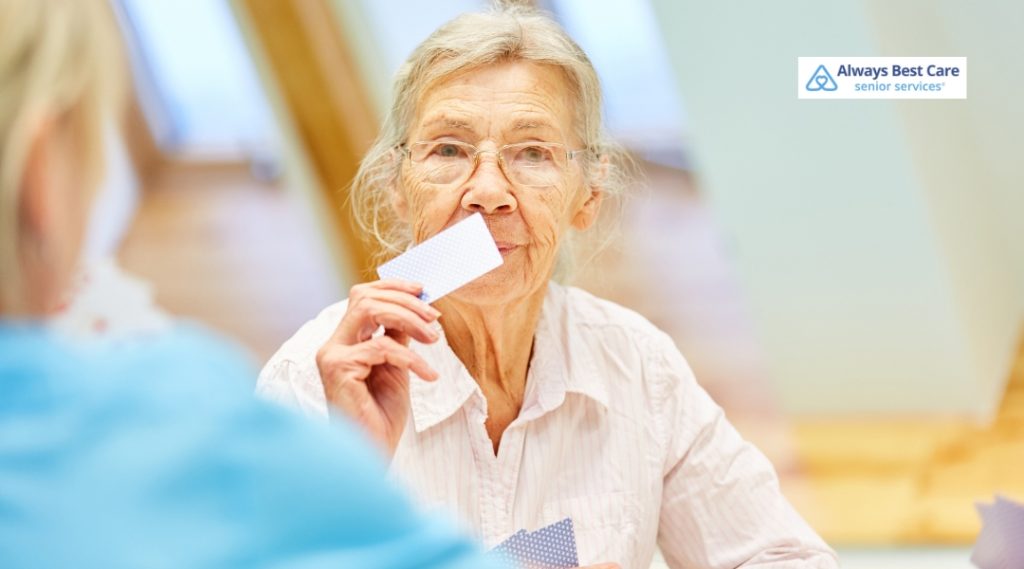 An elderly woman holding a playing card, engaged in a card game with a caregiver, smiling and interacting in a relaxed, warm environment.