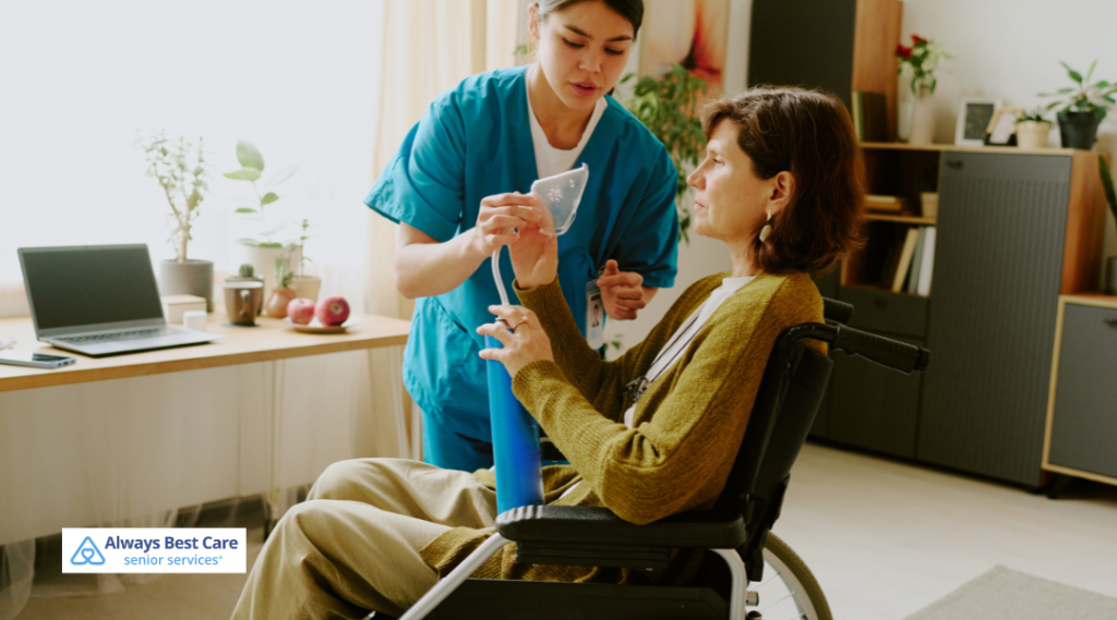 A caregiver in blue scrubs assists a woman in a wheelchair during respiratory therapy, helping her use a breathing device in a bright, organized living space.