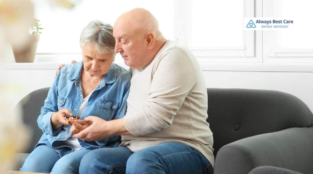A SENIOR COUPLE CHECKS THEIR BLOOD SUGAR LEVELS TOGETHER AT HOME, RECEIVING SUPPORT FROM A CAREGIVER AS THEY MANAGE DIABETES, FOSTERING INDEPENDENCE.