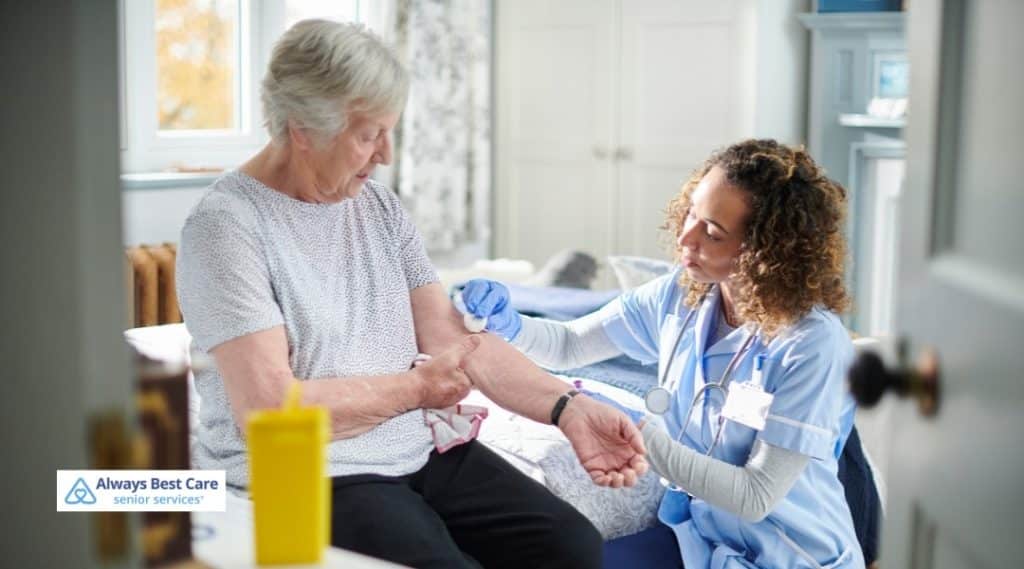 A HEALTHCARE WORKER ADMINISTERS A VACCINE OR TREATMENT TO A SENIOR WOMAN'S ARM WHILE A FAMILY MEMBER OFFERS SUPPORT, SHOWING COMPASSIONATE CARE IN A HOME SETTING.