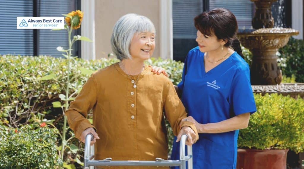 5. CAREGIVER SUPPORTS AN OLDER WOMAN USING A WALKER OUTDOORS IN A GARDEN WALKWAY (ALWAYS BEST CARE LOGO).