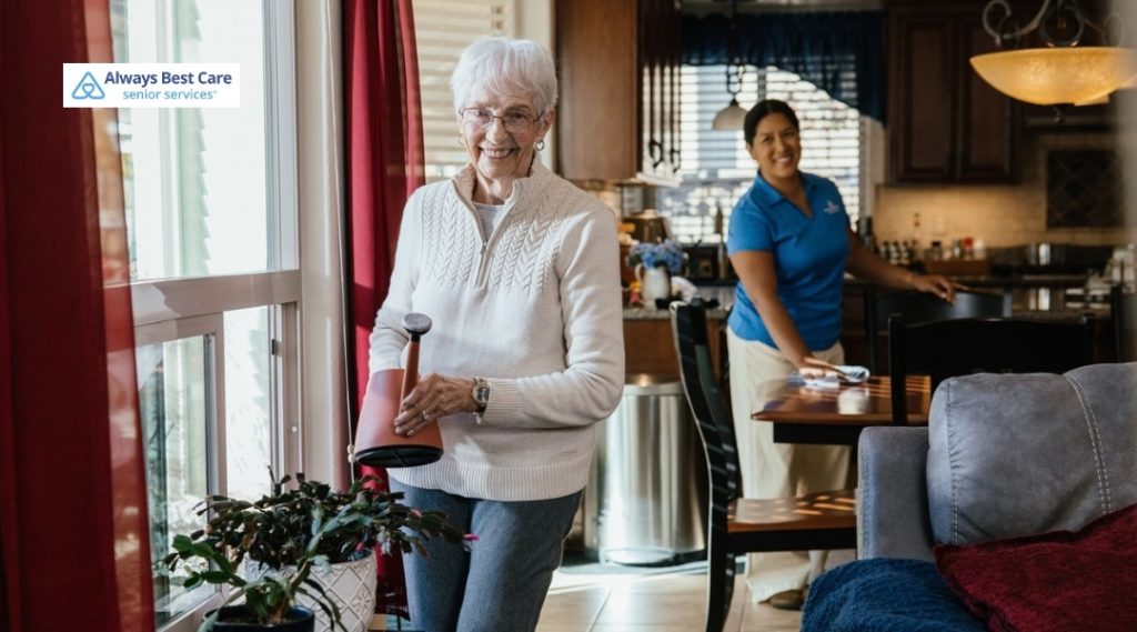 SENIOR WOMAN SMILING WHILE HOLDING A WATERING CAN NEAR A WINDOW AS A CAREGIVER TIDIES THE HOME IN THE BACKGROUND, REPRESENTING INDEPENDENT LIVING SUPPORT.