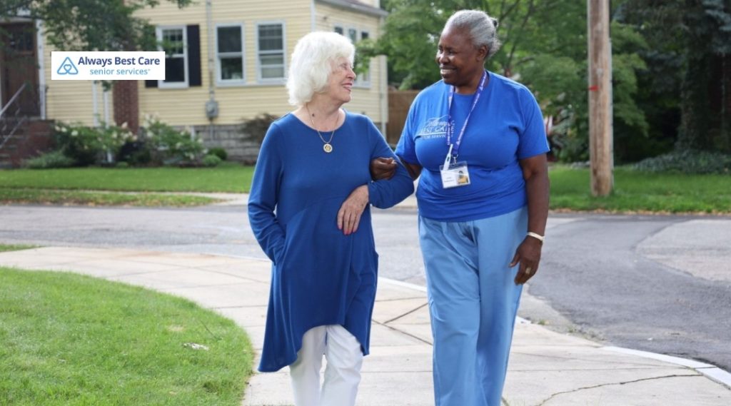 3. CAREGIVER IN BLUE UNIFORM WALKING ARM-IN-ARM WITH AN OLDER WOMAN IN A BLUE TUNIC ALONG A NEIGHBORHOOD SIDEWALK, WITH THE ALWAYS BEST CARE SENIOR SERVICES LOGO IN THE CORNER.
