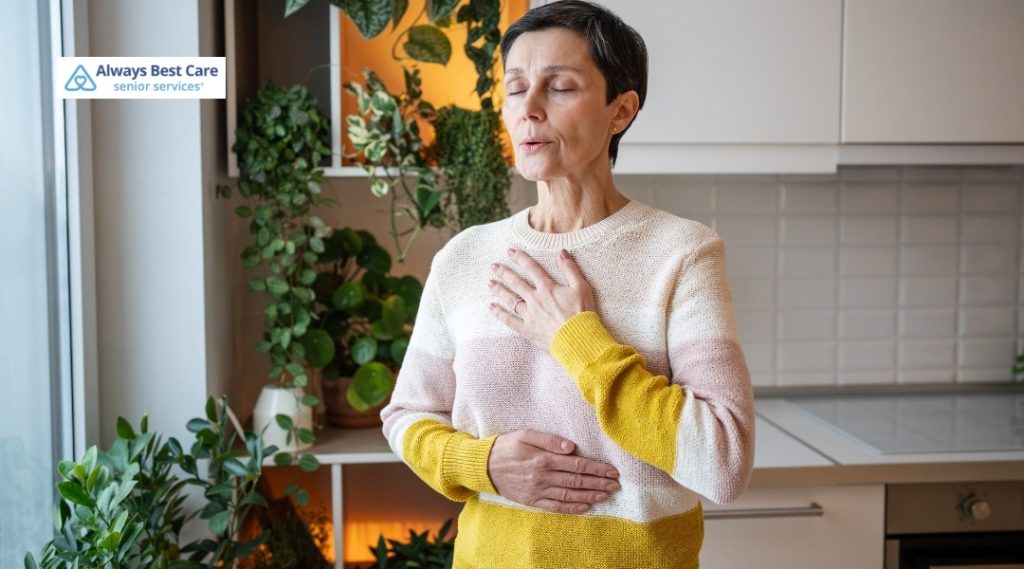 MIDDLE‑AGED WOMAN STANDING INDOORS IN FRONT OF A WALL OF GREEN PLANTS, HOLDING ONE HAND ON HER CHEST AND THE OTHER ON HER STOMACH AS IF EXPERIENCING DISCOMFORT, WITH THE ALWAYS BEST CARE SENIOR SERVICES LOGO IN THE CORNER.