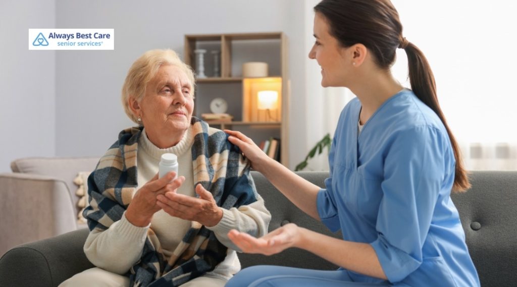 OLDER WOMAN SITTING ON A SOFA HOLDING A PRESCRIPTION PILL BOTTLE WHILE A CAREGIVER IN MEDICAL SCRUBS SITS BESIDE HER, GENTLY TOUCHING HER SHOULDER AND EXPLAINING THE MEDICATION, WITH THE ALWAYS BEST CARE SENIOR SERVICES LOGO IN THE CORNER.
