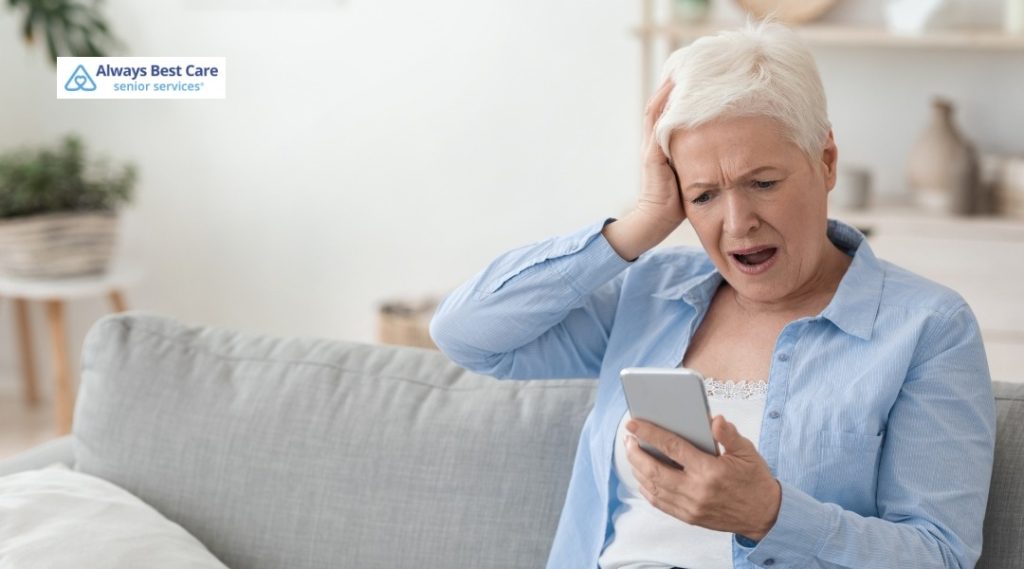 SHOCKED SENIOR WOMAN SITTING ON A SOFA, READING A TROUBLING MESSAGE ON HER SMARTPHONE.