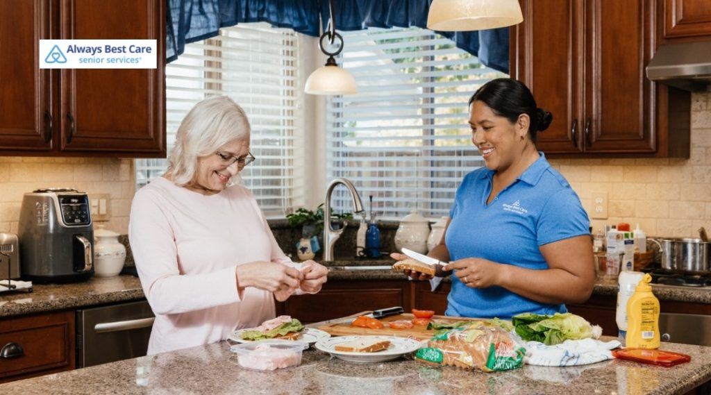 SMILING CAREGIVER AND SENIOR WOMAN PREPARING SANDWICHES TOGETHER IN A BRIGHT KITCHEN, SHOWING COLLABORATIVE MEAL PREP AND COMPANIONSHIP.
