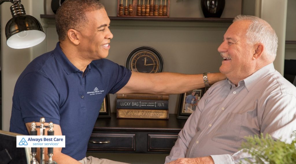A caregiver and a senior man are sharing a lighthearted conversation in a home office setting. The caregiver is gently touching the senior’s shoulder, with both smiling and enjoying the moment.