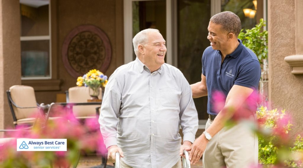A caregiver is assisting a senior man with walking outside on a bright, sunny day. The senior uses a walker, and both share a joyful moment as they walk through a garden.