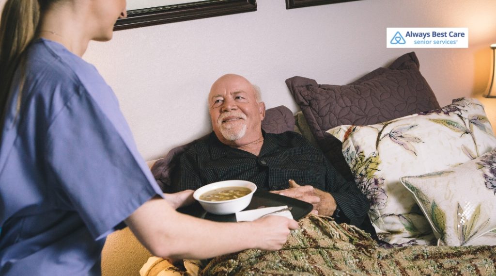 A caregiver is bringing a bowl of soup to a senior man who is comfortably resting in bed. The man smiles at the caregiver, who is offering the food with warmth and care