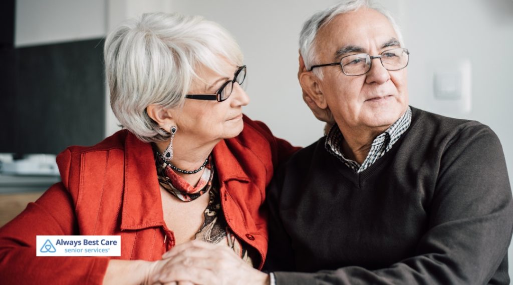 A senior couple sitting together on a couch, smiling and holding hands, looking at each other with warmth and connection. The Always Best Care logo is in the bottom left corner.
