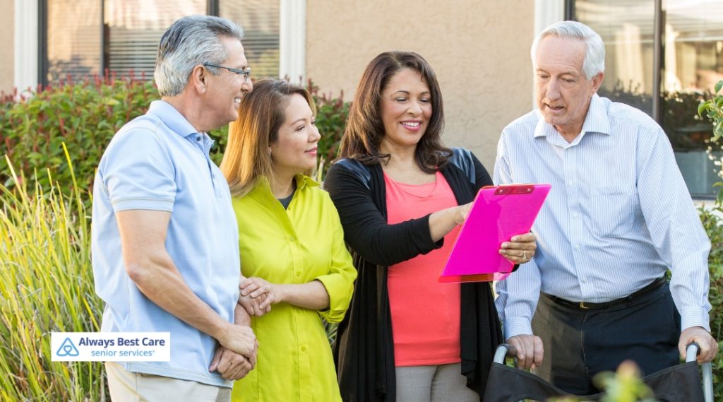 A group of four people, two seniors and two family members, standing together outside, discussing paperwork with a sense of community. The Always Best Care logo is in the bottom left corner.