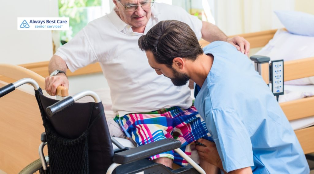 A male caregiver helps an elderly man transfer from a bed to a wheelchair in a home care setting. The scene emphasizes safety, mobility assistance, and supportive caregiving. Always Best Care branding is shown.