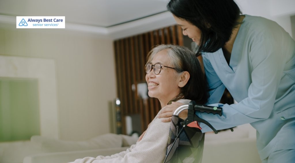 A caregiver in scrubs stands behind an older woman in a wheelchair, offering a reassuring touch on her shoulders. The woman smiles, reflecting warmth, comfort, and professional care. The Always Best Care logo is visible.