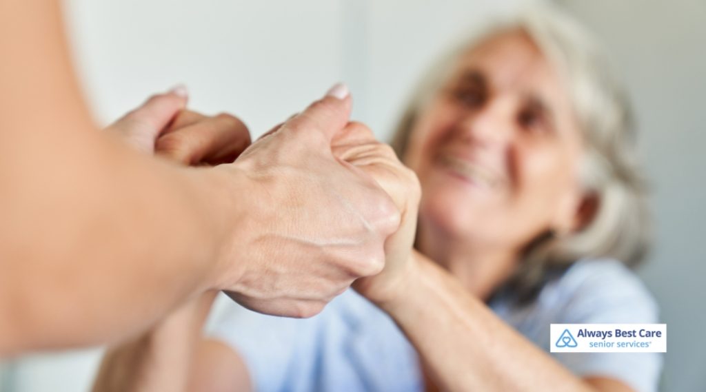 A close-up image showing a caregiver gently holding hands with a smiling senior woman in the background. The focus on handholding highlights emotional support, compassion, and trust. The Always Best Care logo is displayed.