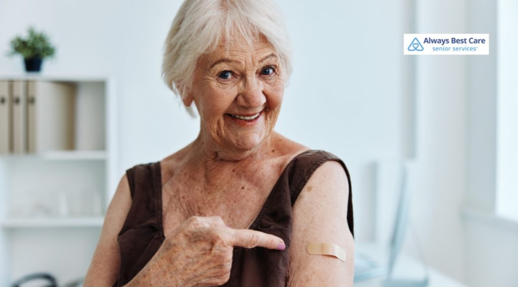 A senior woman with gray hair, smiling while pointing at a band-aid on her arm after receiving a vaccine shot.