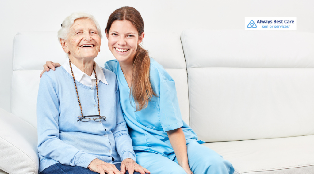 A senior woman smiling with a caregiver by her side, both enjoying a warm and friendly moment on a bright, comfortable sofa.