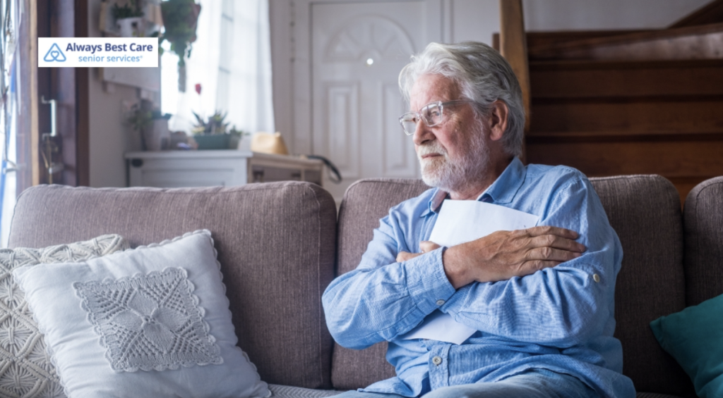 A senior man sitting alone on a sofa, looking pensive and holding a piece of paper. The room around him is bright and cozy, but he appears reflective and somewhat distant.