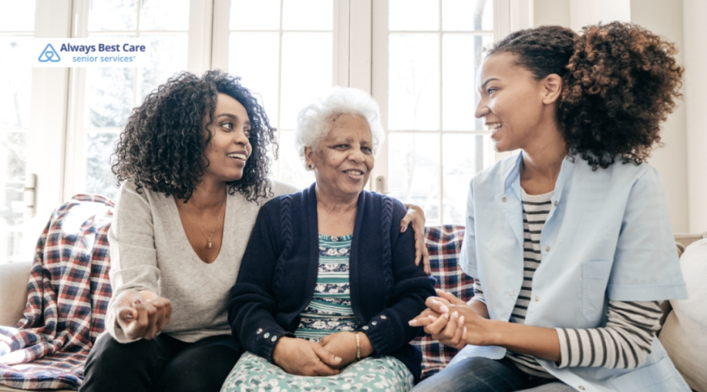 A family photo with a senior woman seated in the middle, smiling as she is surrounded by her adult daughter and a caregiver. They are in a bright and inviting indoor space.