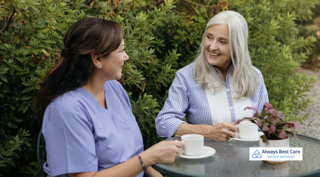 Two women, one elderly and one caregiver, sitting together at a table outdoors, smiling as they enjoy a cup of tea. The setting is serene and green, with plants around them.