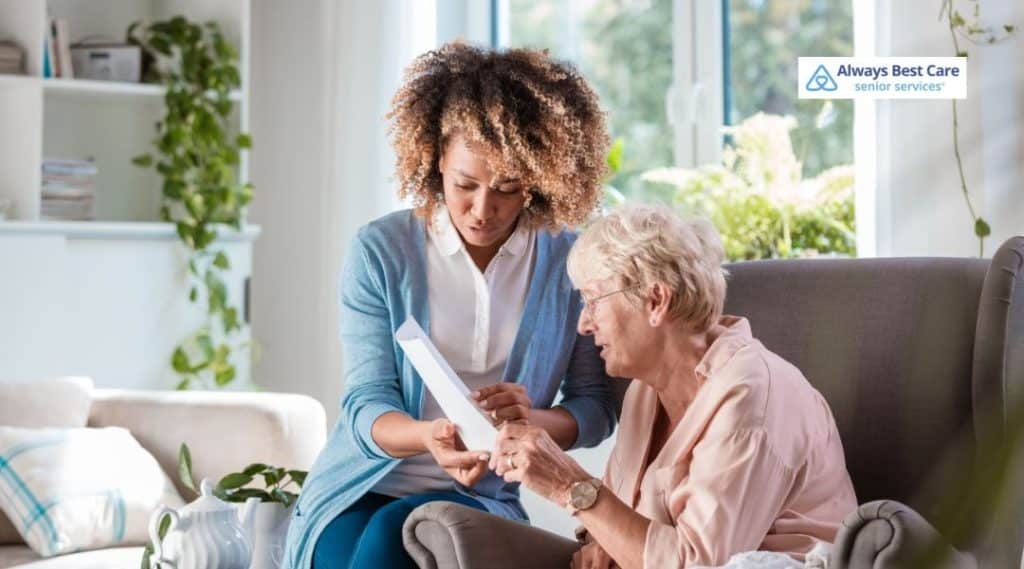 Caregiver sitting beside an older adult at home, gently reviewing paperwork and providing guidance in a calm, supportive setting.