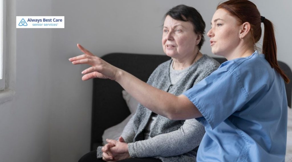 Nurse in blue scrubs pointing toward a window while explaining something to an older woman seated beside her.