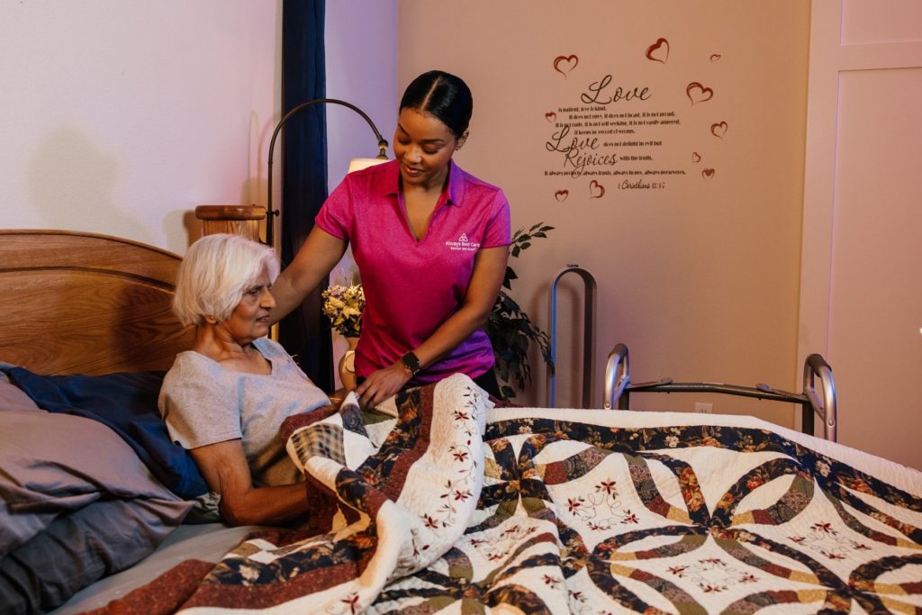 Caregiver in a pink Always Best Care polo gently adjusting the blanket of an older woman sitting up in bed, in a softly lit bedroom decorated with a quilt and a wall quote about love.