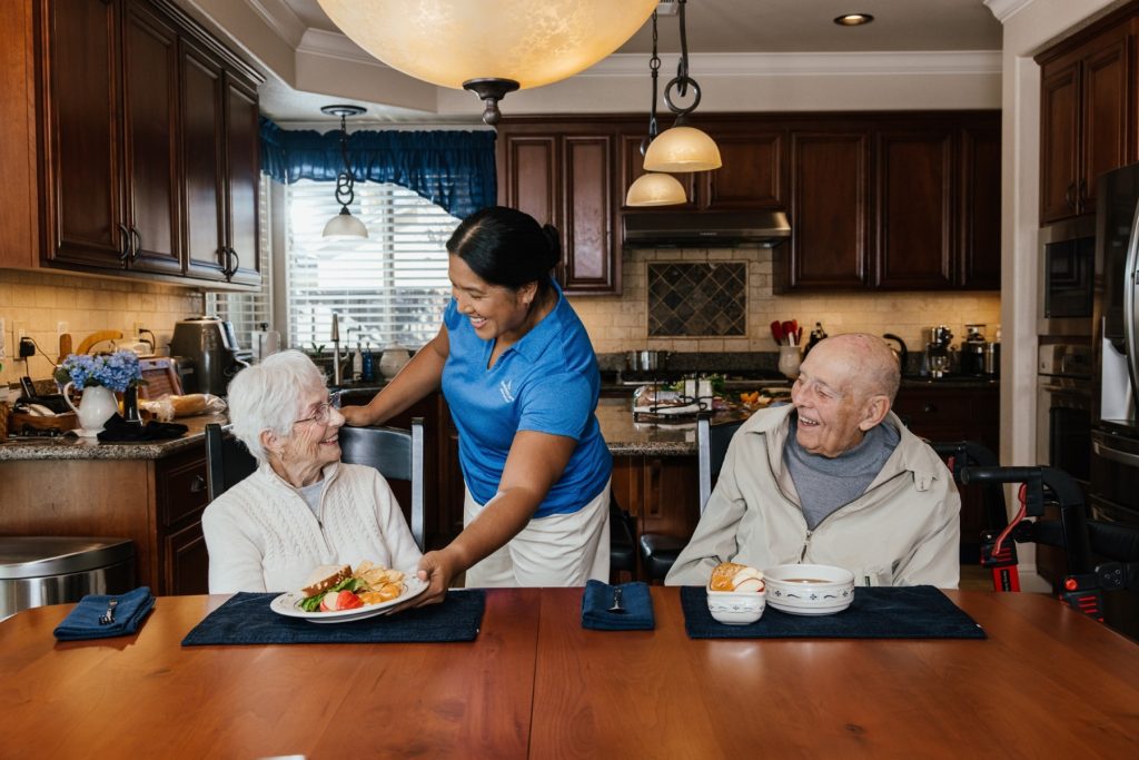 Caregiver serves a meal to an older couple seated at their kitchen table; the man has a walker beside him.