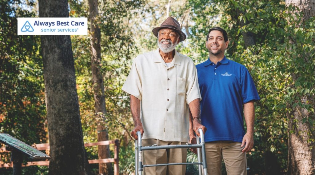An older man using a walker in an outdoor setting, accompanied by a caregiver who stands beside him offering support. Both are smiling, showing a positive and caring interaction. The caregiver wears a blue shirt with the "Always Best Care" logo.