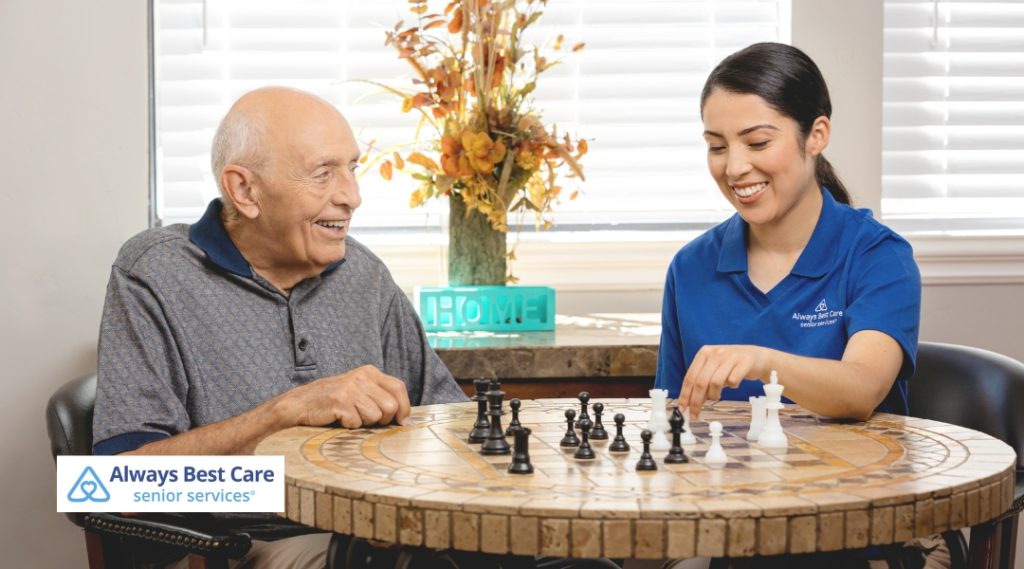 An elderly man and his caregiver engaged in a friendly game of chess at a table. The man is smiling, and the caregiver is focused on the game. The caregiver wears a blue "Always Best Care" shirt. The setting is indoor with natural light coming from the window.