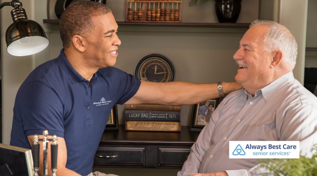 A caregiver and an elderly man sharing a joyful moment in a home office setting. The caregiver, dressed in a blue shirt, places a supportive hand on the man’s shoulder as they smile at each other. The background includes books and a clock, creating a warm, homey atmosphere.