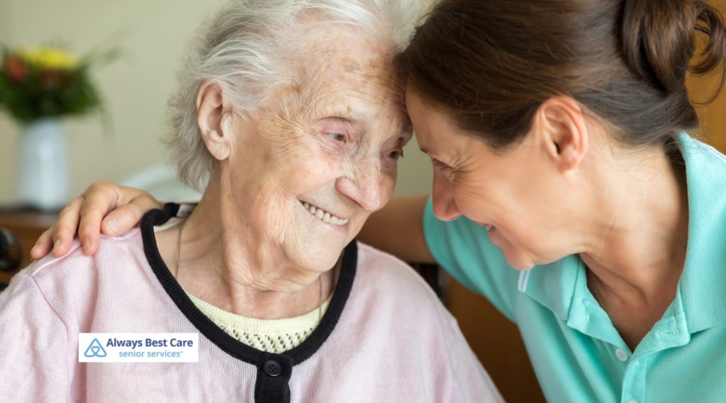 A caregiver and an elderly woman share a heartfelt moment, smiling and leaning close to each other, with a background of soft, natural lighting and greenery, highlighting the emotional connection between them.